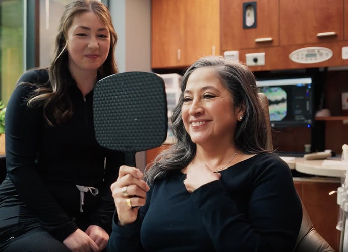 A woman is holding hand-mirror and smiling at her dentist